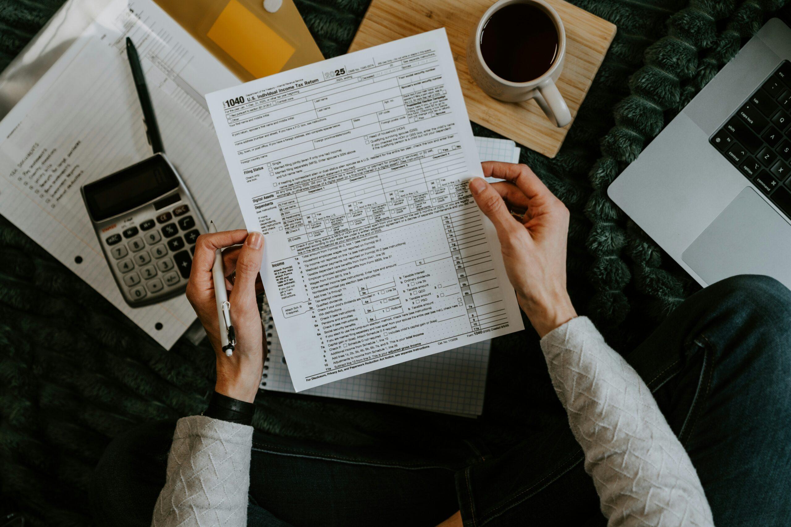 Person holding and reviewing a 1040 U.S. tax form with a pen, surrounded by a calculator, notebook, coffee mug, and laptop on a cozy setup.